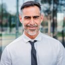 Vertical portrait of successful mature caucasian business man smiling looking at camera standing outdoors at workplace with cheerful attitude. Front view of handsome executive employee wearing a suit. High quality photo